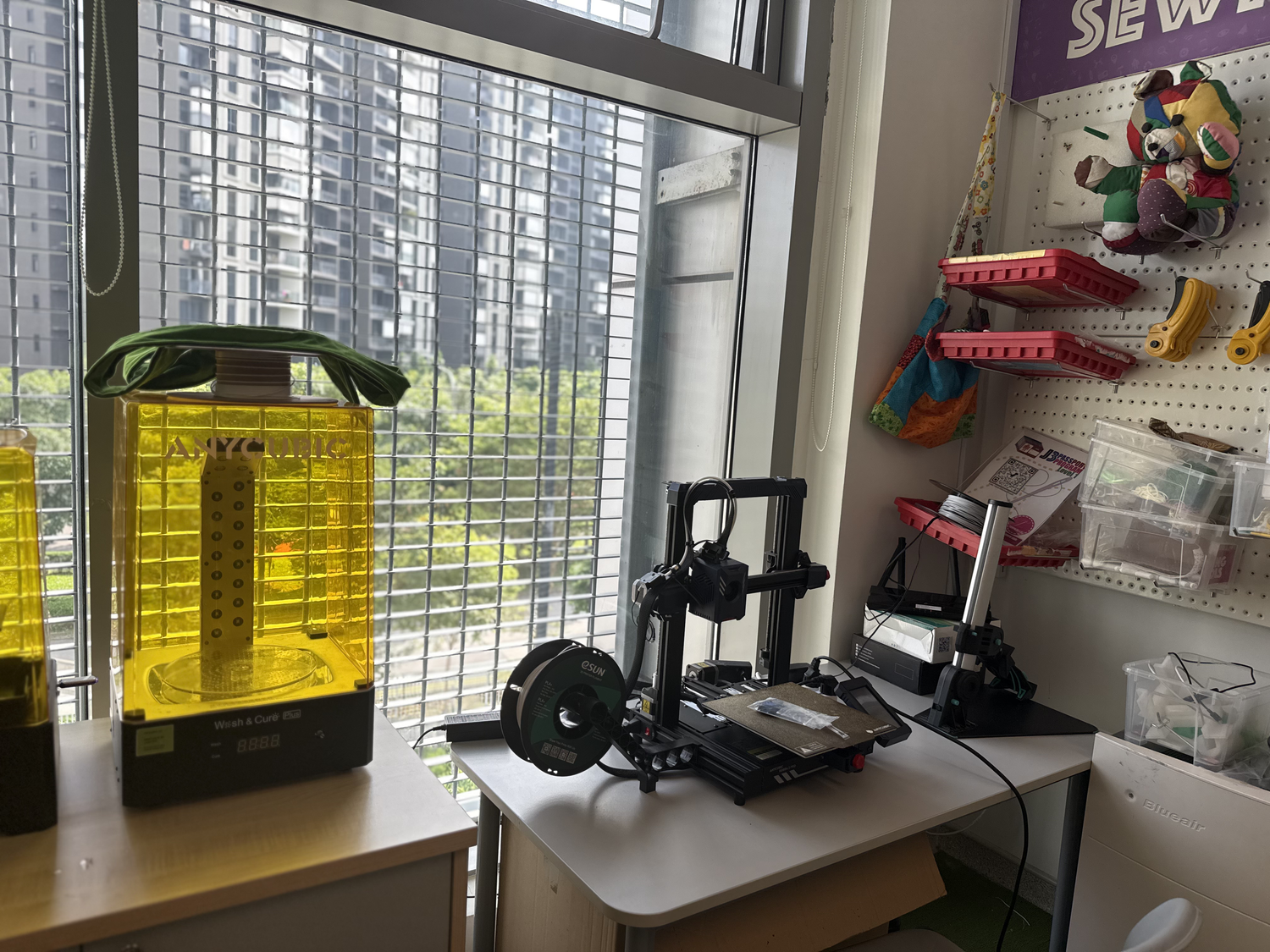 窓辺の机に置かれた3Dプリンターと、壁のボードに布や工具が並ぶ部屋