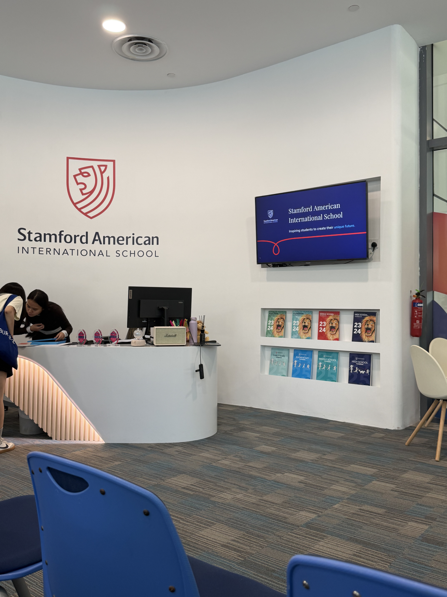 Stamford American International Schoolのロゴと校名が掲げられた白い壁の受付エリア、右側には校名を映すディスプレイと年刊誌が並ぶ棚