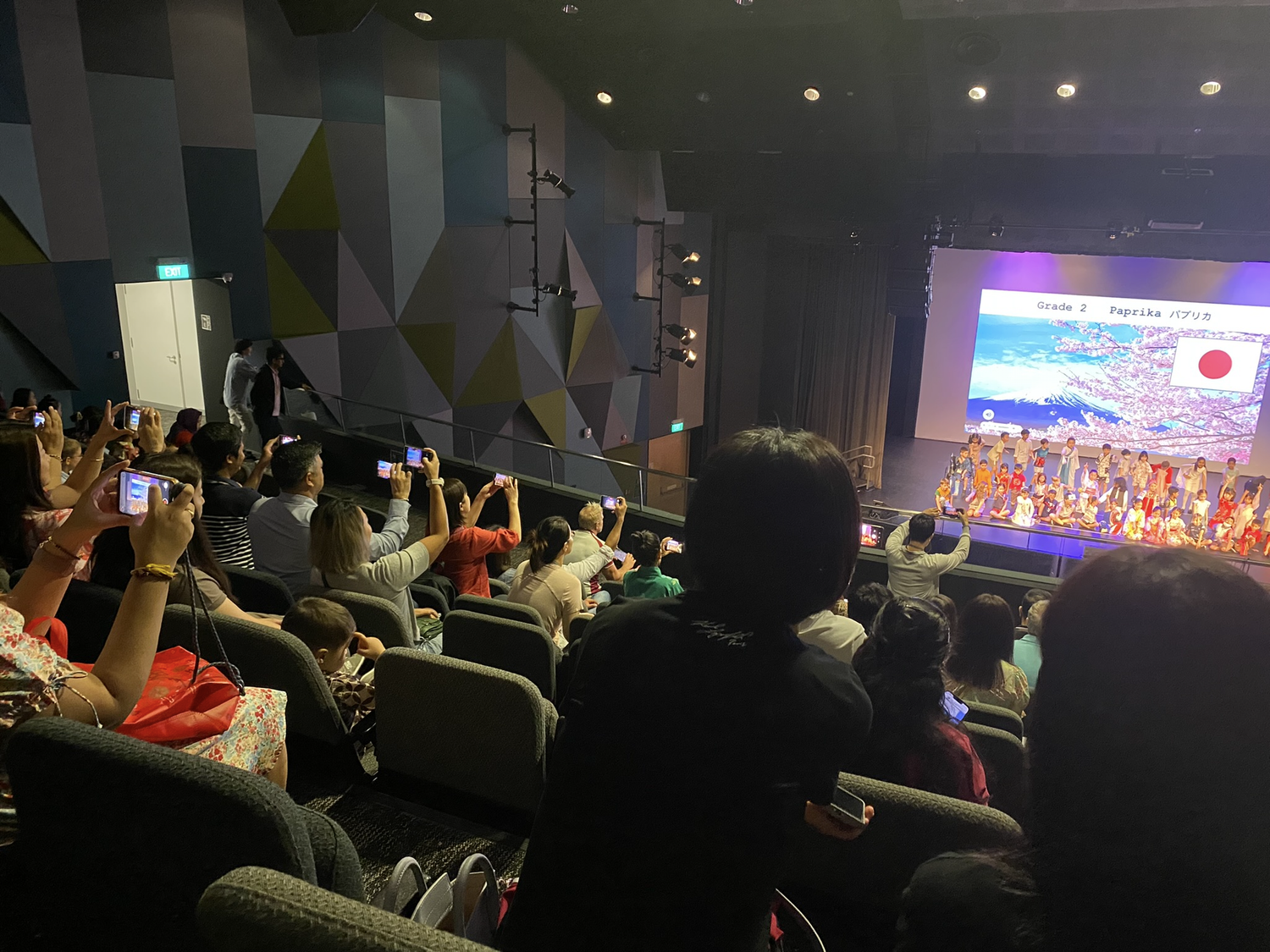 客席から舞台に向けてスマートフォンを構える人たちと、日本の国旗が映るスクリーン
